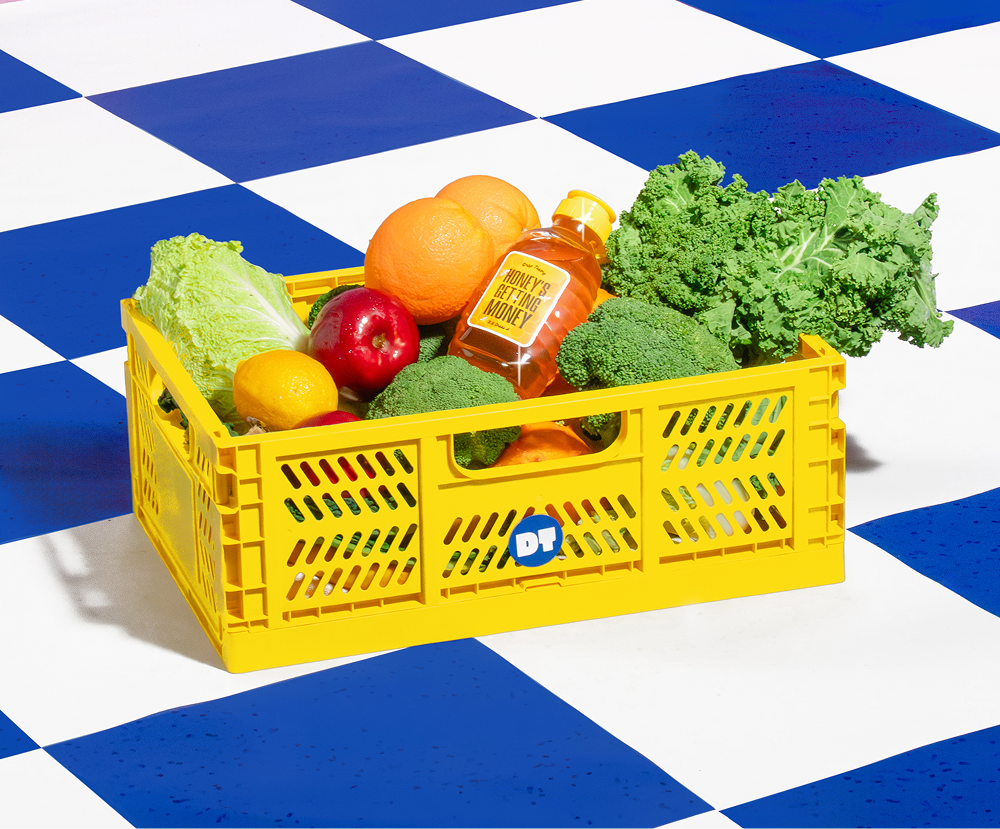 Yellow crate filled with fresh produce including kale, broccoli, oranges, apple, lemon, lettuce, and a bottle of honey on a blue and white checkered floor.