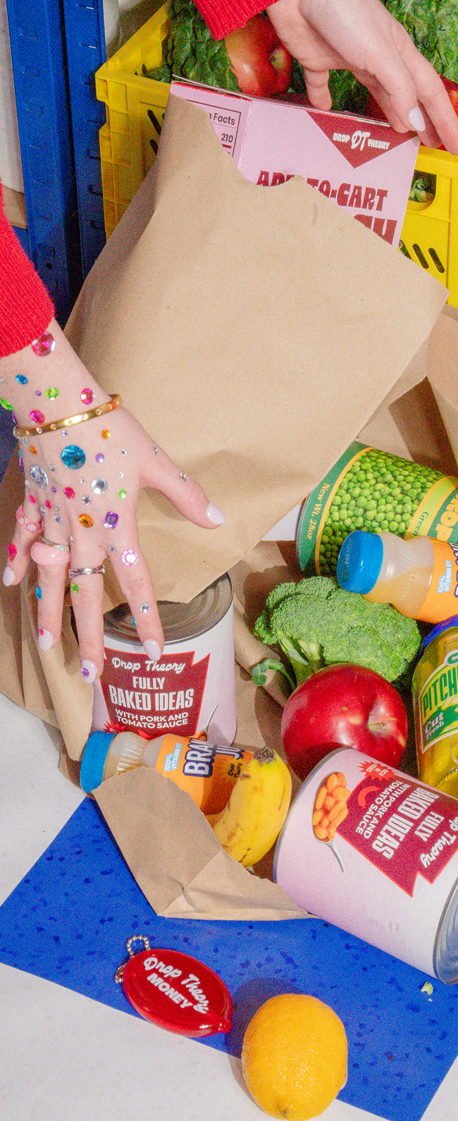 Hands with colorful rhinestone gems place grocery items including canned beans, produce, and packaged goods into a brown paper bag from a yellow crate.