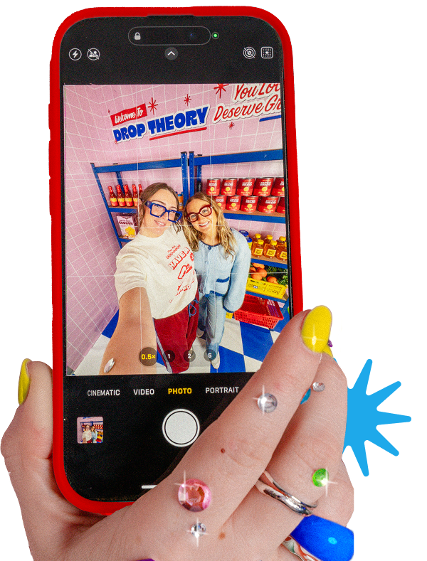 Hand holding a smartphone taking a selfie of two people in a colorful “Drop Theory” grocery-style set with shelves of products in the background.